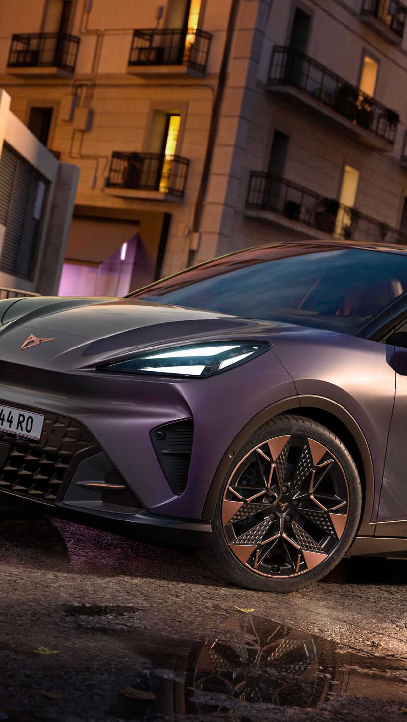 Vůz CUPRA Raval v barvě Plasma se střechou Manhattan Grey stojí v moderním industriálním prostředí. Přední tříčtvrteční pohled zvýrazňuje ostré linie karoserie, měděná aerodynamická kola a osvětlené logo CUPRA. V pozadí je výrazný nápis „Raval“, který podtrhuje identitu tohoto elektrického městského vozu.
